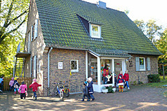 Ein zweistöckiges, helles Haus mit grünem Dach. Vor dem Haus stehen mehrere Kinder, die miteinander spielen. 