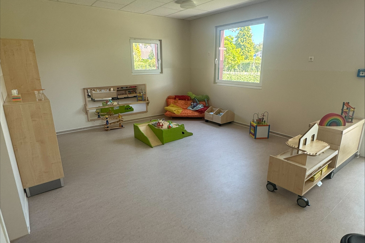 Ein heller und geräumiger Spielraum mit verschiedenen Spielmöglichkeiten. Es gibt Teppiche, Spielzeuge und Möbel wie Regale und Tische. Fenster lassen Tageslicht herein, und einige Sitzbereiche mit Kissen sind sichtbar. Der Boden ist hell und einladend.