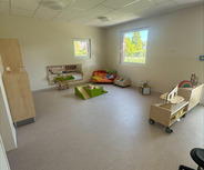 Ein heller und geräumiger Spielraum mit verschiedenen Spielmöglichkeiten. Es gibt Teppiche, Spielzeuge und Möbel wie Regale und Tische. Fenster lassen Tageslicht herein, und einige Sitzbereiche mit Kissen sind sichtbar. Der Boden ist hell und einladend.