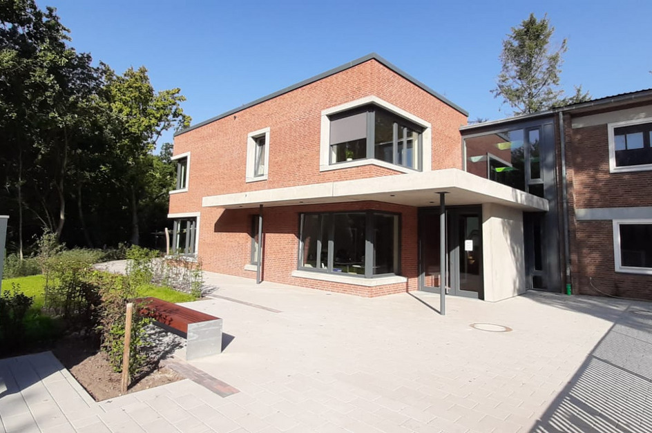 Ein modernes, zweigeschossiges Gebäude aus roten Ziegeln mit großen Fenstern und einem überdachten Eingangsbereich. 