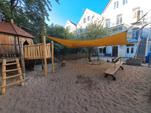 Spielplatz mit einem Holzspielhaus, einer Rutsche und einer Holzschaukel. Ein gelbes Sonnensegel hängt über einem sandigen Untergrund. Im Hintergrund sind mehrere Gebäude und ein paar Bäume sichtbar.