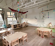 Ein heller, geräumiger Kita-Raum mit mehreren Holztischen und Stühlen. Dekorative Elemente hängen von der Decke, darunter bunte Papiergirlanden. An der Wand befindet sich ein Kalender und Spielmaterialien. Ein kleines Spielhaus steht in der Ecke, und große Fenster lassen Tageslicht herein.