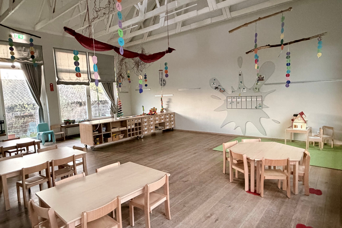 Ein heller, geräumiger Kita-Raum mit mehreren Holztischen und Stühlen. Dekorative Elemente hängen von der Decke, darunter bunte Papiergirlanden. An der Wand befindet sich ein Kalender und Spielmaterialien. Ein kleines Spielhaus steht in der Ecke, und große Fenster lassen Tageslicht herein.