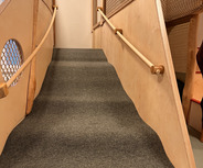 Eine Innenansicht einer Treppe mit einem weichen, grauen Teppichbelag. Die Treppe führt zu einer Spielplattform, umgeben von holz- und netzartigen Strukturen. Die Wände sind hell, und es gibt ein Fenster, das Tageslicht hereinlässt.