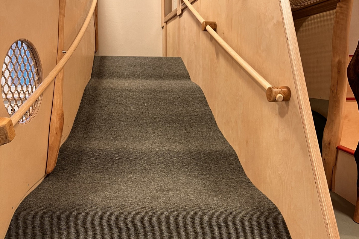 Eine Innenansicht einer Treppe mit einem weichen, grauen Teppichbelag. Die Treppe führt zu einer Spielplattform, umgeben von holz- und netzartigen Strukturen. Die Wände sind hell, und es gibt ein Fenster, das Tageslicht hereinlässt.