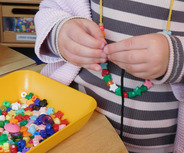Ein Kind fädelt bunte Perlen auf eine Schnur. Eine gelbe Schale mit weiteren Perlen in verschiedenen Farben und Formen steht daneben.