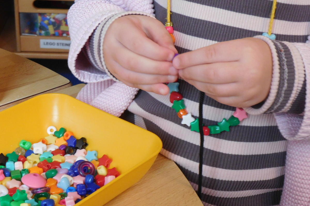 Ein Kind fädelt bunte Perlen auf eine Schnur. Eine gelbe Schale mit weiteren Perlen in verschiedenen Farben und Formen steht daneben.