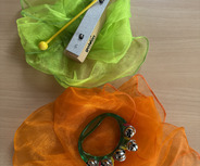 Auf einem Tisch liegt ein grüner und ein orangefarbener, transparenter Stoff. Auf dem grünen Stoff befindet sich ein Holz-Xylophon mit einem gelben Schlägel. Auf dem orangefarbenen Stoff liegt ein Tamburin mit mehreren klingenden Schellen.