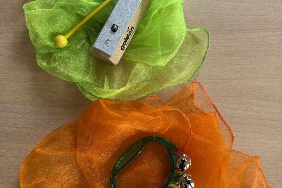 Auf einem Tisch liegt ein grüner und ein orangefarbener, transparenter Stoff. Auf dem grünen Stoff befindet sich ein Holz-Xylophon mit einem gelben Schlägel. Auf dem orangefarbenen Stoff liegt ein Tamburin mit mehreren klingenden Schellen.