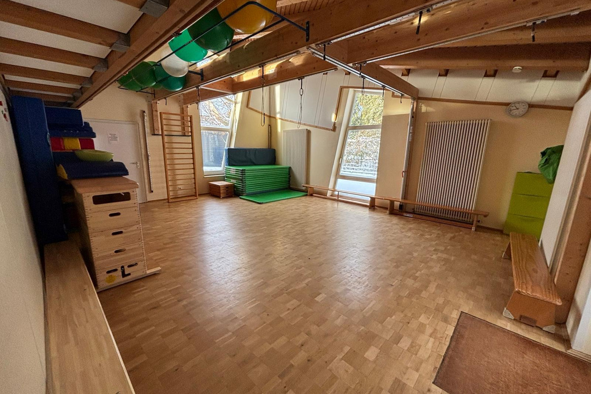 Der Raum ist hell und geräumig, mit Holzboden und großen Fenstern. An einer Wand stehen bunten Turnmatten und Geräte. In der Mitte befindet sich eine grüne Trainingsmatte. Eine Wand hat ein Klettergerüst und an der Decke hängen verschiedene Bälle.