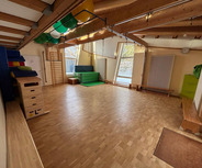 Der Raum ist hell und geräumig, mit Holzboden und großen Fenstern. An einer Wand stehen bunten Turnmatten und Geräte. In der Mitte befindet sich eine grüne Trainingsmatte. Eine Wand hat ein Klettergerüst und an der Decke hängen verschiedene Bälle.