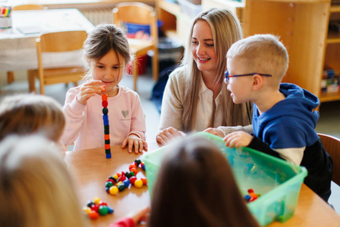 Ev. Kindertagesstätte "Kinder Garten Eden" Gemünden. Blick auf einen Tisch mit spielenden Kindern. Ein Mädchen sitzt neben einer Erzieherin und spielt mit bunten Steinen. Im unscharfen Vordergrund zwei weitere Kinder.