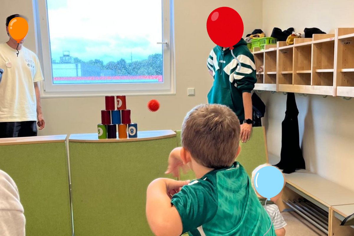 Ein kleines Kind wirft einen Ball auf einen Turm aus bunten Bechern. Im Hintergrund sind zwei Erwachsene zu sehen, einer davon trägt eine grüne Jacke. Der Raum hat Fenster, die Tageslicht hereinlassen, und ist mit einfachen Möbeln ausgestattet.