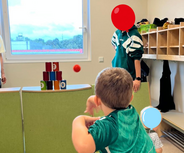 Ein kleines Kind wirft einen Ball auf einen Turm aus bunten Bechern. Im Hintergrund sind zwei Erwachsene zu sehen, einer davon trägt eine grüne Jacke. Der Raum hat Fenster, die Tageslicht hereinlassen, und ist mit einfachen Möbeln ausgestattet.