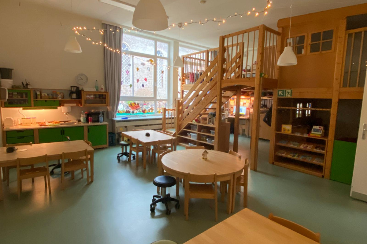 Ein heller Raum mit mehreren Tischgruppen und Stühlen, eine Küche in grünem Design und ein Holzspielbereich mit einer Treppe in der Ecke. 