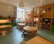 Ein heller Raum mit mehreren Tischgruppen und Stühlen, eine Küche in grünem Design und ein Holzspielbereich mit einer Treppe in der Ecke. 