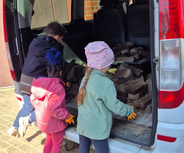 Drei Kinder stehen am offenen Kofferraum eines Lieferwagens und heben Steine oder Platten hinein. Sie tragen Handschuhe und scheinen beschäftigt zu sein.