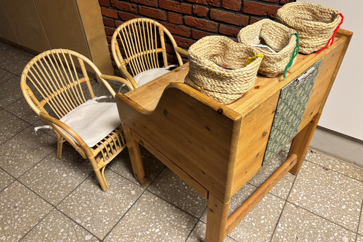 Im Bild sind zwei kleine Rattanstühle neben einem kleinen Tresen aus Holz zu sehen. Auf dem Tisch befinden sich drei geflochtene Körbe in verschiedenen Farben. 