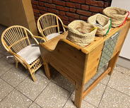 Im Bild sind zwei kleine Rattanstühle neben einem kleinen Tresen aus Holz zu sehen. Auf dem Tisch befinden sich drei geflochtene Körbe in verschiedenen Farben. 