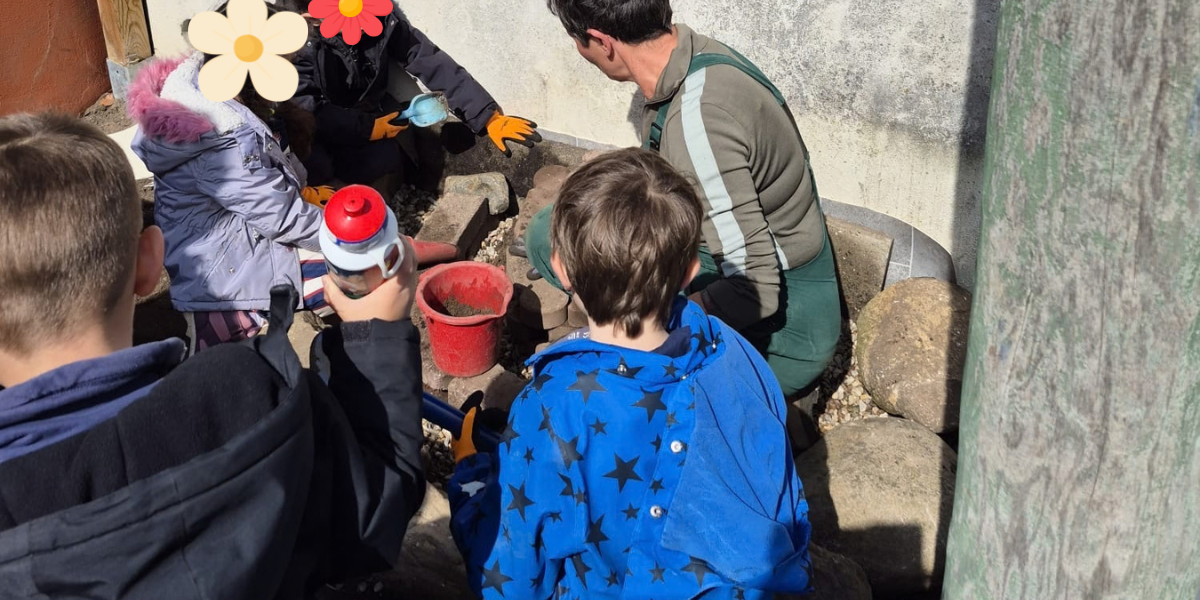 Eine Gruppe von Kindern und einer Erwachsenen arbeitet im Freien. Die Erwachsene zeigt den Kindern, wie man mit Werkzeugen und Materialien umgeht. Im Hintergrund sind Steine und Eimer sichtbar. Es ist sonnig und die Kinder tragen einfache Outdoor-Kleidung.