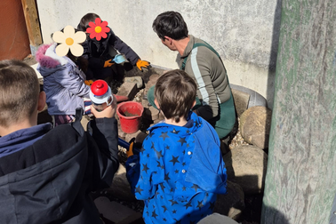 Eine Gruppe von Kindern und einer Erwachsenen arbeitet im Freien. Die Erwachsene zeigt den Kindern, wie man mit Werkzeugen und Materialien umgeht. Im Hintergrund sind Steine und Eimer sichtbar. Es ist sonnig und die Kinder tragen einfache Outdoor-Kleidung.
