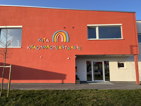 Die Bildbeschreibung zeigt die Vorderseite eines Gebäudes in leuchtendem Rot. Ein bunter Schriftzug „Kita Krachmacherstraße“ ist gut sichtbar über dem Eingang angebracht. Es gibt Fenster an der Fassade und eine kleine Grünfläche im Vordergrund.