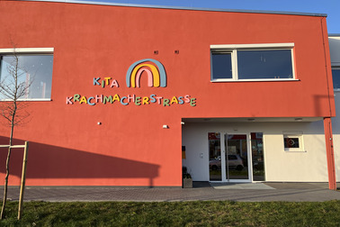 Die Bildbeschreibung zeigt die Vorderseite eines Gebäudes in leuchtendem Rot. Ein bunter Schriftzug „Kita Krachmacherstraße“ ist gut sichtbar über dem Eingang angebracht. Es gibt Fenster an der Fassade und eine kleine Grünfläche im Vordergrund.