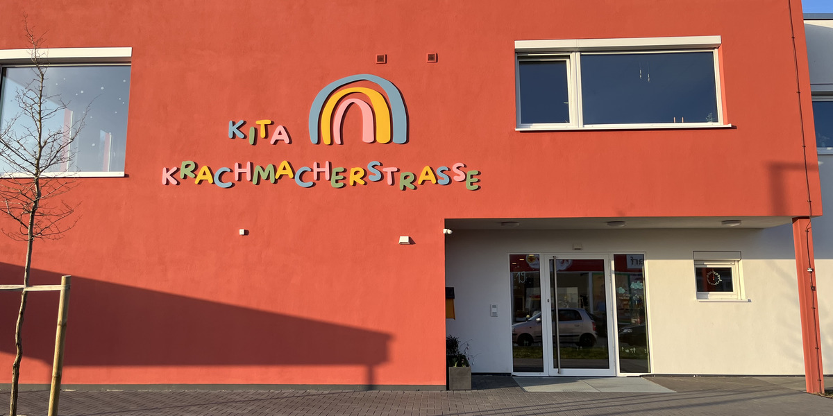 Die Bildbeschreibung zeigt die Vorderseite eines Gebäudes in leuchtendem Rot. Ein bunter Schriftzug „Kita Krachmacherstraße“ ist gut sichtbar über dem Eingang angebracht. Es gibt Fenster an der Fassade und eine kleine Grünfläche im Vordergrund.