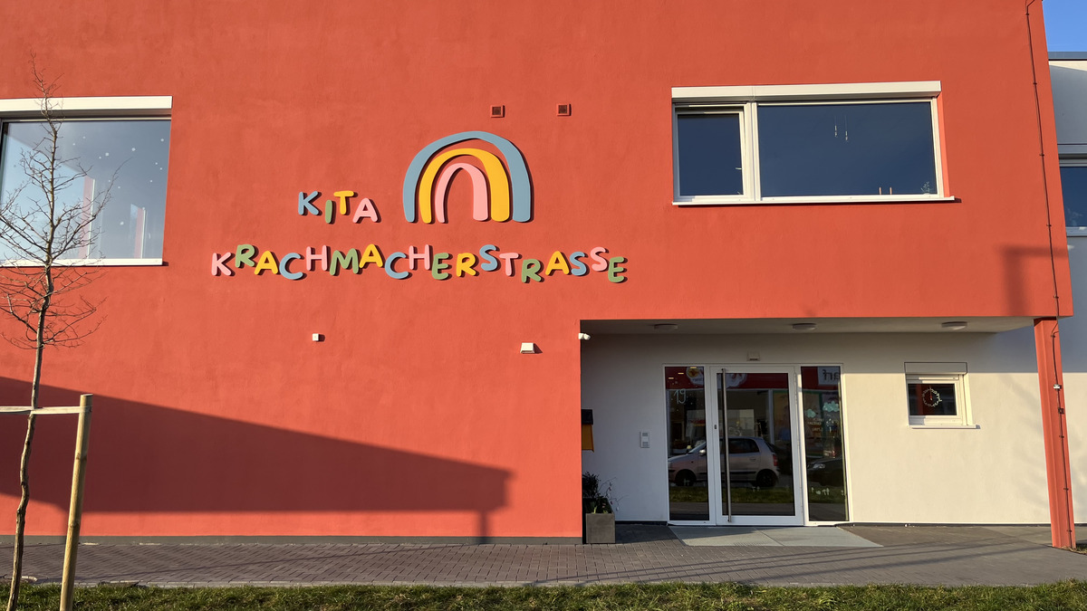 Die Bildbeschreibung zeigt die Vorderseite eines Gebäudes in leuchtendem Rot. Ein bunter Schriftzug „Kita Krachmacherstraße“ ist gut sichtbar über dem Eingang angebracht. Es gibt Fenster an der Fassade und eine kleine Grünfläche im Vordergrund.