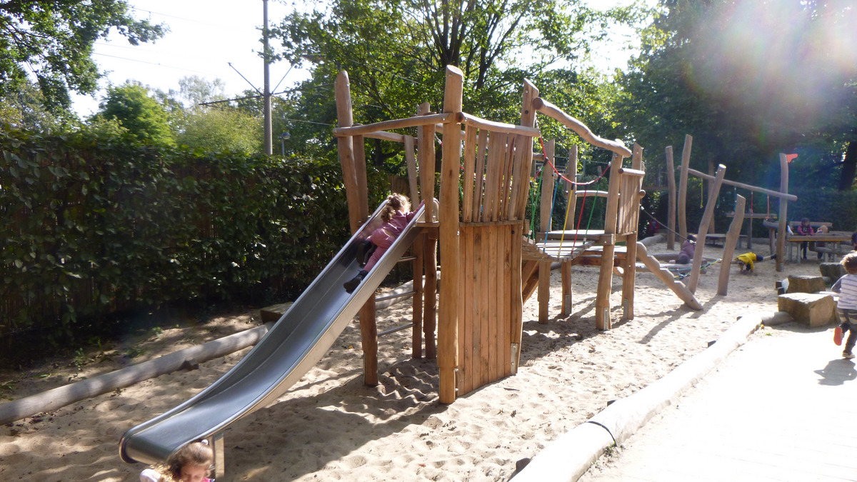 Ein Spielplatz mit einer Holzkonstruktion, die eine Rutsche umfasst. Ein Kind rutscht die Rutsche hinunter. 