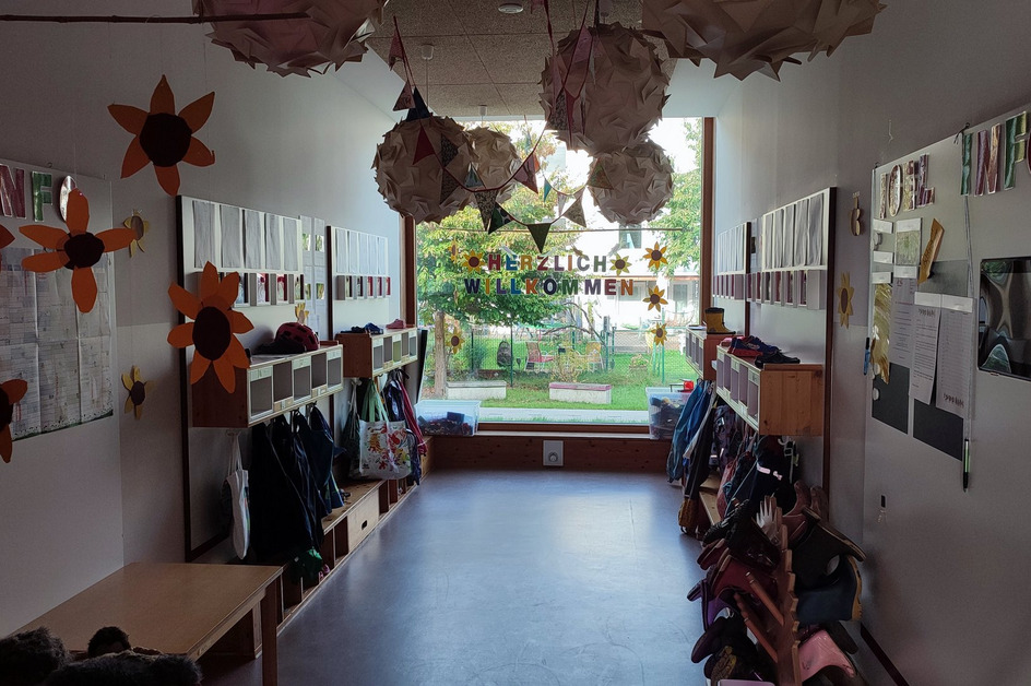 Ein einladender Flur mit hängenden Papiersachen und einer großen Fensterscheibe, durch die ein Garten zu sehen ist. An der Wand steht "HERZLICH WILLKOMMEN". Es gibt mehrere Garderoben mit Kinderjacken und Taschen. 