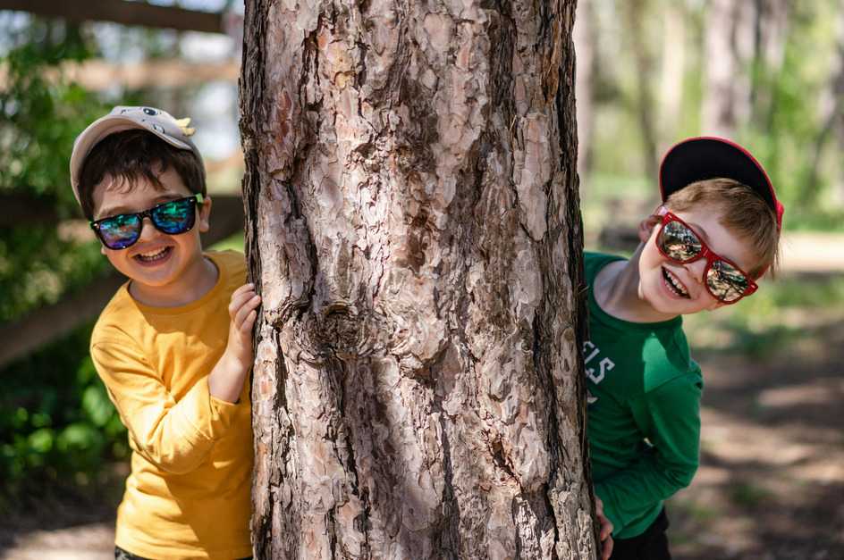 Zwei fröhliche Kinder verstecken sich hinter einem großen Baum. Der Junge links trägt ein gelbes Shirt und Sonnenbrille, während der Junge rechts ein grünes Shirt und eine rote Cap hat. Beide lächeln in die Kamera und scheinen Spaß zu haben.