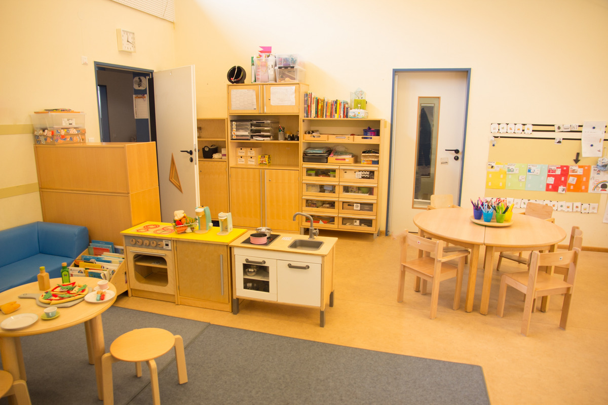 Ein heller, freundlicher Kindergartenraum mit einer blauen Couch, einem Spieltisch mit Spielzeug und einer kleinen Spielküche. An einer Wand hängen bunte Zeichnungen und ein Regal mit Büchern ist sichtbar. Ein runder Tisch mit Stühlen bietet Platz zum Spielen und Arbeiten.