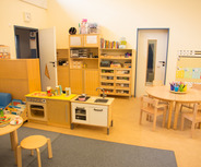 Ein heller, freundlicher Kindergartenraum mit einer blauen Couch, einem Spieltisch mit Spielzeug und einer kleinen Spielküche. An einer Wand hängen bunte Zeichnungen und ein Regal mit Büchern ist sichtbar. Ein runder Tisch mit Stühlen bietet Platz zum Spielen und Arbeiten.