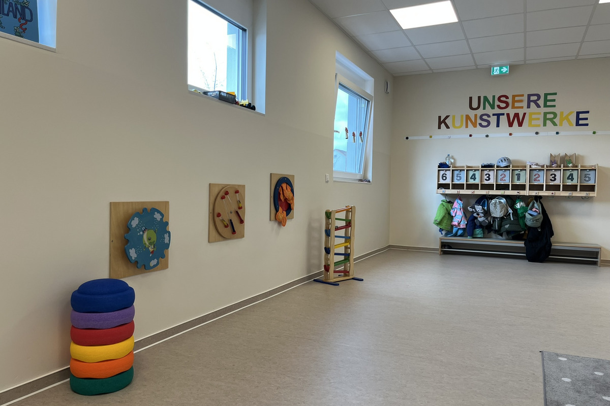 Ein heller Raum mit bunten Wandspielzeugen und einer Aufhängemöglichkeit für Jacken. An der Wand steht "UNSERE KUNSTWERKE" in bunten Buchstaben. In einer Ecke sind farbige, gestapelte Kissen sichtbar.