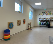 Ein heller Raum mit bunten Wandspielzeugen und einer Aufhängemöglichkeit für Jacken. An der Wand steht "UNSERE KUNSTWERKE" in bunten Buchstaben. In einer Ecke sind farbige, gestapelte Kissen sichtbar.