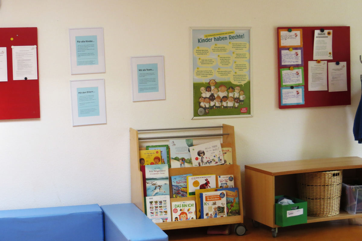 Der Raum zeigt eine Bücherwand mit einer Vielzahl von Kinderbüchern. An der Wand hängen Informationsblätter und ein Plakat mit dem Titel „Kinder haben Rechte!“, das wichtige Rechte von Kindern hervorhebt. Ein blauer Sitzbereich ergänzt die Einrichtung.