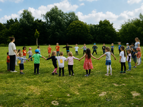 Eine Gruppe von Kindern steht im Freien auf einer Wiese und hält sich an den Händen. Sie bilden einen Kreis und scheinen gemeinsam zu spielen oder zu singen. Im Hintergrund sind Bäume und blauer Himmel zu sehen.