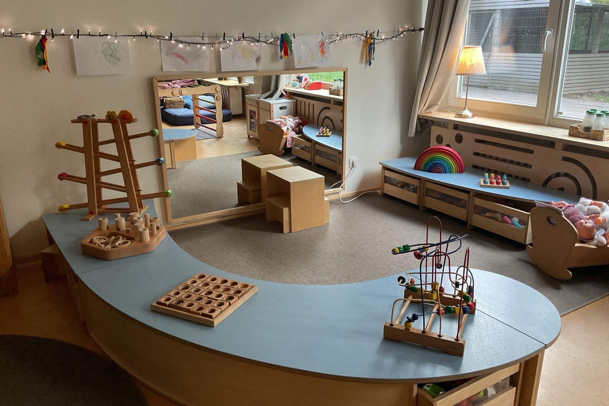 Ein Krippenraum mit Spielzeugen. Im Vordergrund stehen mehrere Holzwürfel und eine Kugelbahn. An den Wänden hängen Zeichnungen, und ein großer Spiegel reflektiert den Raum. 