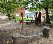 Ein Spielplatz mit Holzspielgeräten, darunter eine Kletterlandschaft aus natürlichen Materialien und eine große runde Holzfigur. Die Umgebung ist grün mit Bäumen und im Hintergrund ist der Kindergarten zu sehen.