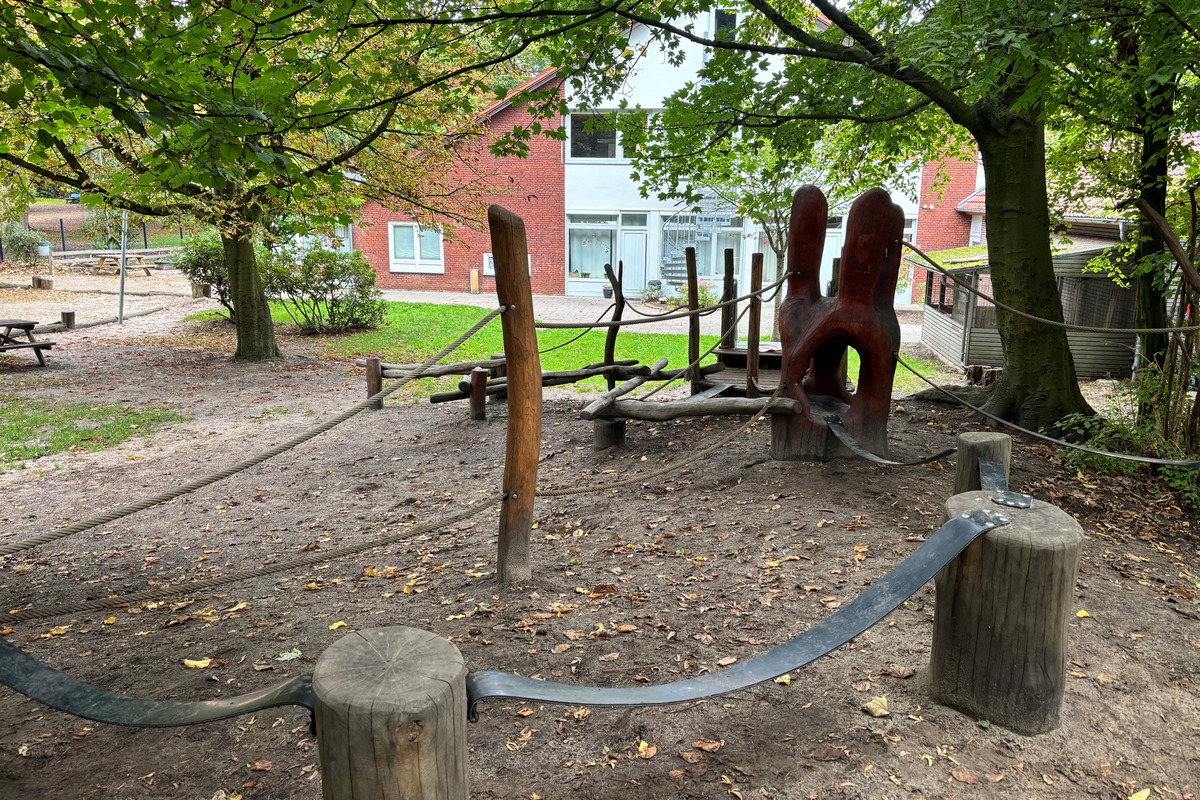 Ein Spielplatz mit Holzspielgeräten, darunter eine Kletterlandschaft aus natürlichen Materialien und eine große runde Holzfigur. Die Umgebung ist grün mit Bäumen und im Hintergrund ist der Kindergarten zu sehen.