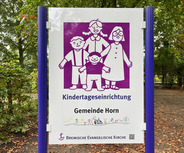 Ein Schild einer Kindertageseinrichtung zeigt eine bunte Illustration von einer Familie mit zwei Erwachsenen und drei Kindern. Darunter steht der Text "Kindertageseinrichtung Gemeinde Horn" und das Logo der Bremischen Evangelischen Kirche. 
