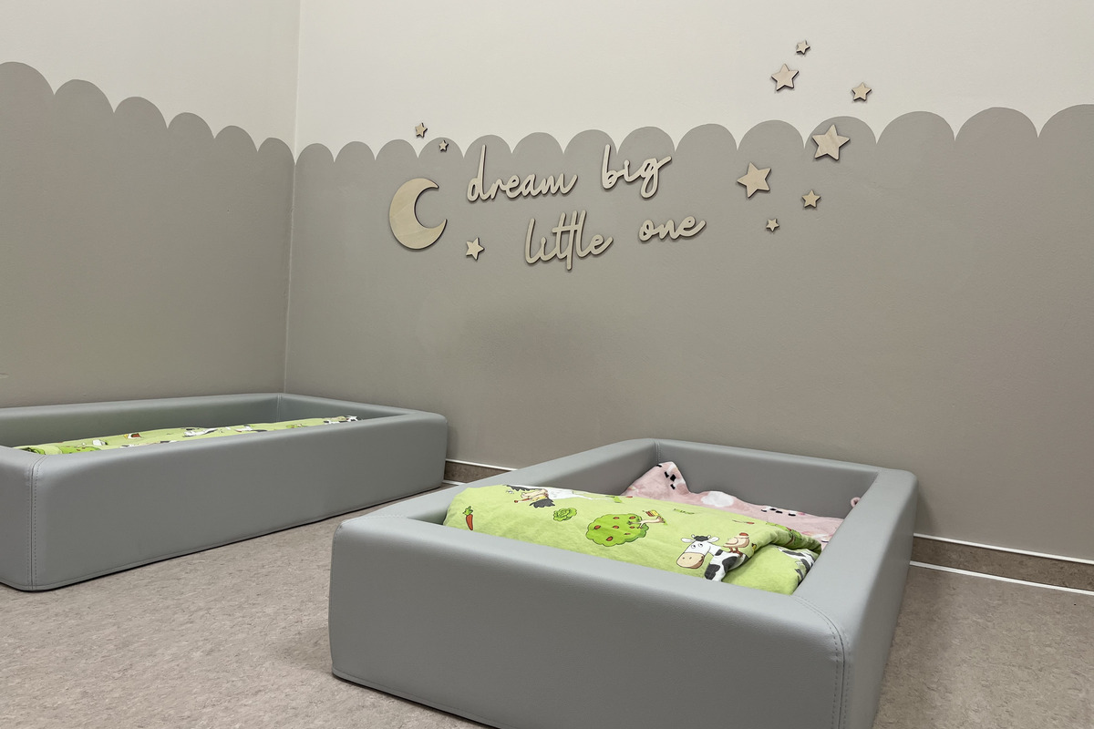 Ein gemütlicher Raum mit zwei grauen Betten, die mit bunten Decken ausgestattet sind. An der Wand steht der Schriftzug „dream big little one“ über einer dekorativen Halbmond- und Sternenmotivik.