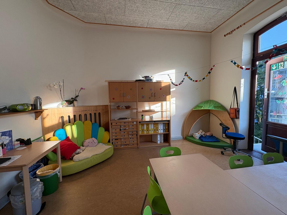 Der Raum ist eine helle, freundliche Einrichtung für Kinder. Es gibt eine Sitzecke mit bunten Kissen, einen Spielbereich mit einem grünen Zelt und einen Tisch mit Stühlen. An den Wänden hängen dekorative Girlanden. Ein Fenster bietet Tageslicht und einen Blick nach draußen.