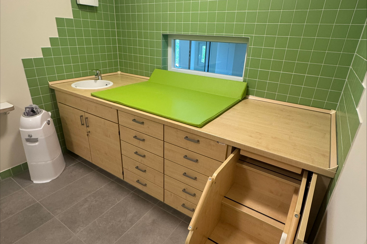 Ein Raum mit grünen Fliesenwänden, einem Wickeltisch aus Holz und einem Waschbecken. Auf der Konsole liegt eine grüne Wickelauflage. Rechts befindet sich eine Holztreppe. 