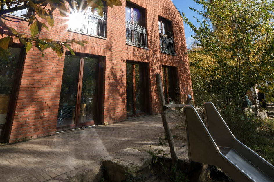 Ein rotes Backsteingebäude mit großen Fensterfronten steht im Vordergrund. Umgeben von herbstlichen Bäumen mit bunten Blättern. Im Vordergrund ist eine Rutsche aus Metall zu sehen. Der Himmel ist blau mit einigen Wolken.
