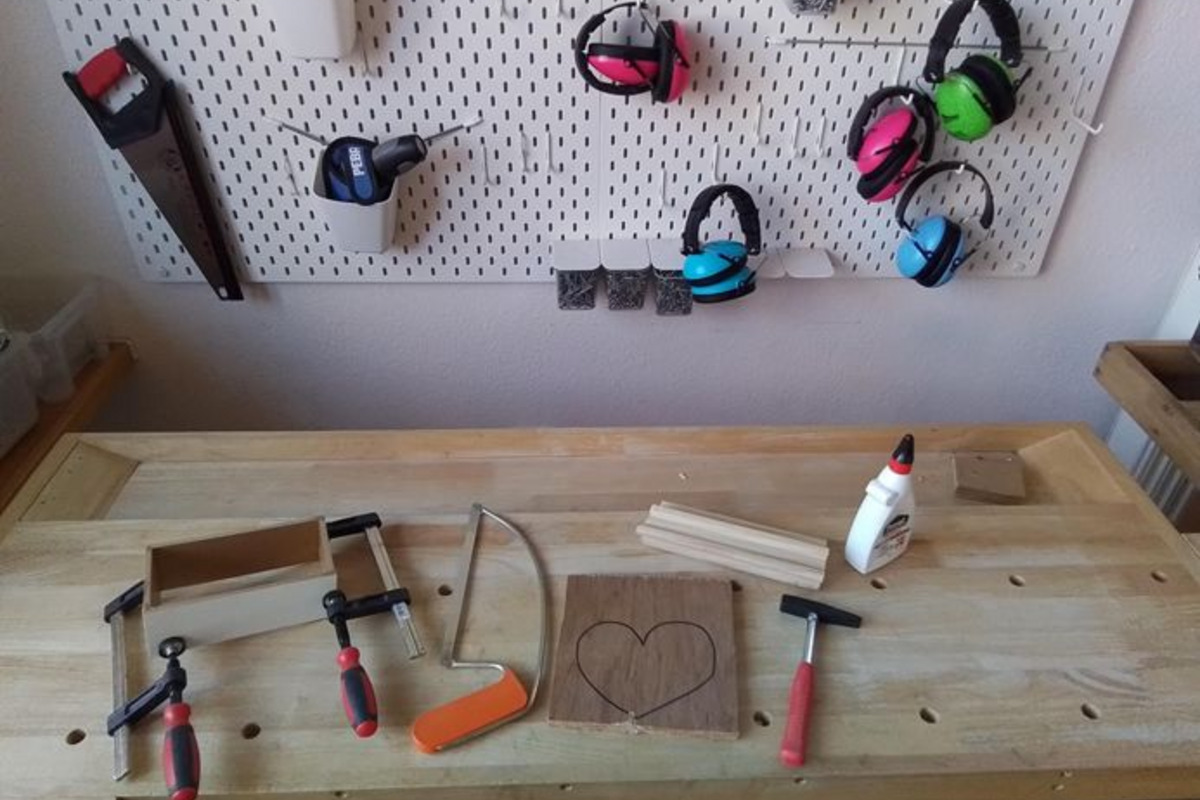 Auf einem Tisch liegen verschiedene Werkzeug- und Bastelutensilien. Ein Holzteil zeigt eine Herzform, daneben sind Klemmen, ein Sägeblatt, Leim und Holzlatten angeordnet. An der Wand hängen Ohrenschützer in verschiedenen Farben.