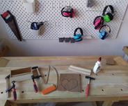 Auf einem Tisch liegen verschiedene Werkzeug- und Bastelutensilien. Ein Holzteil zeigt eine Herzform, daneben sind Klemmen, ein Sägeblatt, Leim und Holzlatten angeordnet. An der Wand hängen Ohrenschützer in verschiedenen Farben.