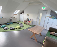 Ein heller Raum mit schrägen Fenstern und einem weichen grünen Teppich. An den Wänden stehen Bücherregale mit einer Auswahl an Kinderbüchern. Auf dem Boden liegen bunte Kissen zum Sitzen. Ein Holzschreibtisch und ein bequemer Sessel sind ebenfalls vorhanden.