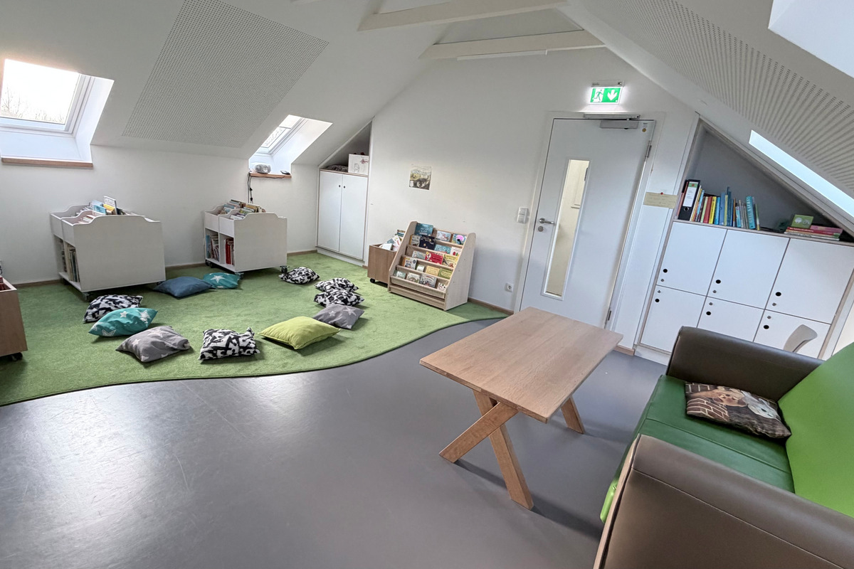 Ein heller Raum mit schrägen Fenstern und einem weichen grünen Teppich. An den Wänden stehen Bücherregale mit einer Auswahl an Kinderbüchern. Auf dem Boden liegen bunte Kissen zum Sitzen. Ein Holzschreibtisch und ein bequemer Sessel sind ebenfalls vorhanden.